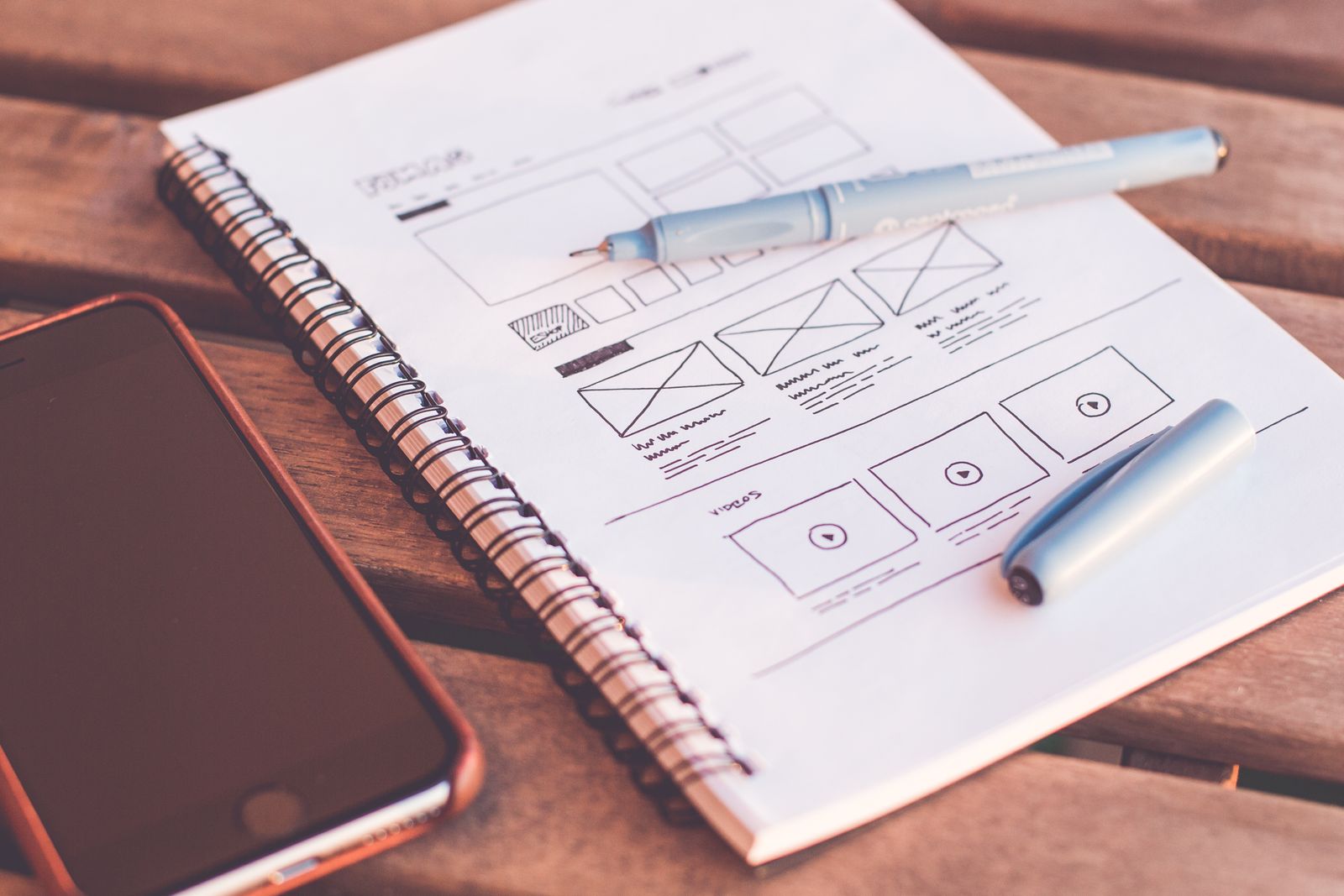 A notebook with hand-drawn website wireframes, two pens and a phone on a wooden desk